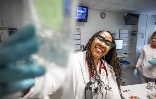 A smiling Cleveland Clinic nurse wearing glasses and a stethoscope adjusts an IV bag in a hospital unit.