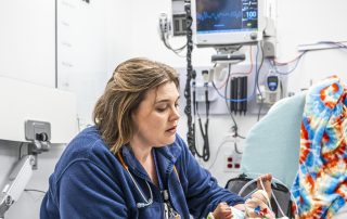 A woman nurse wearing a stethoscope holds an infant patient in a clinical setting with medical monitoring equipment behind them.