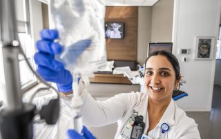 A smiling Cleveland Clinic nurse wearing gloves adjusts an IV fluid bag in a hospital room.