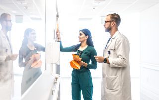 A Cleveland Clinic nurse in green scrubs writes on a whiteboard while collaborating with a doctor in a white coat in a hospital hallway.