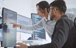 A woman points at code on a computer monitor while collaborating with a male colleague in an office setting with multiple screens displaying programming.
