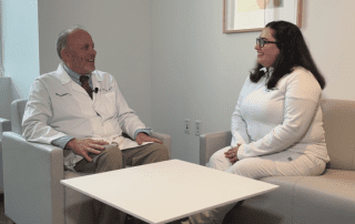 A male doctor and a female nurse, both in white coats, sit across from each other in a lounge area and share a conversation.