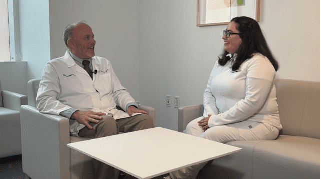 A male doctor and a female nurse, both in white coats, sit across from each other in a lounge area and share a conversation.