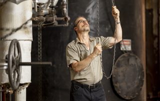 A man wearing glasses and a work shirt pulls on a heavy chain while inspecting industrial pipes and valves in a mechanical room.