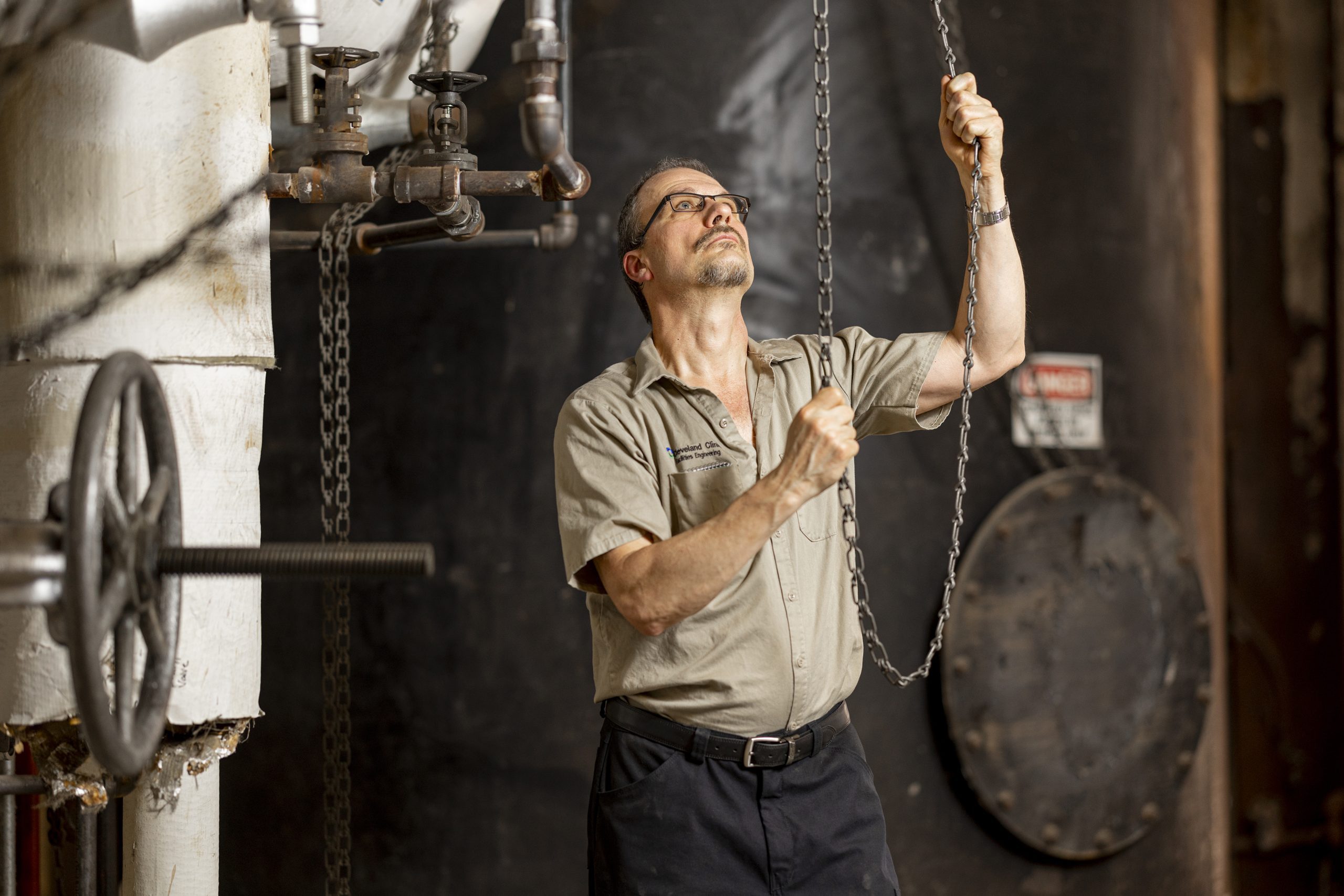 A man wearing glasses and a work shirt pulls on a heavy chain while inspecting industrial pipes and valves in a mechanical room.