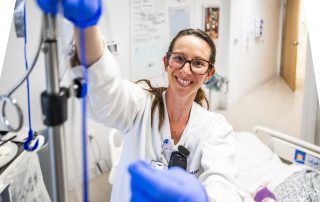 A smiling Cleveland Clinic nurse wearing glasses and gloves adjusts IV tubing beside a patient’s hospital bed.