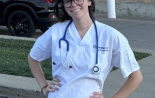 A Cleveland Clinic employee, Gabby Repko, in white scrubs with a stethoscope around her neck smiles while standing outdoors.