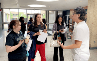 A Cleveland Clinic employee in white scrubs speaks with a small group of people holding papers and drinks in a lobby area.