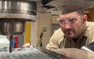 A man wearing safety glasses operates a large industrial milling machine, closely monitoring the drill bit cutting into metal.