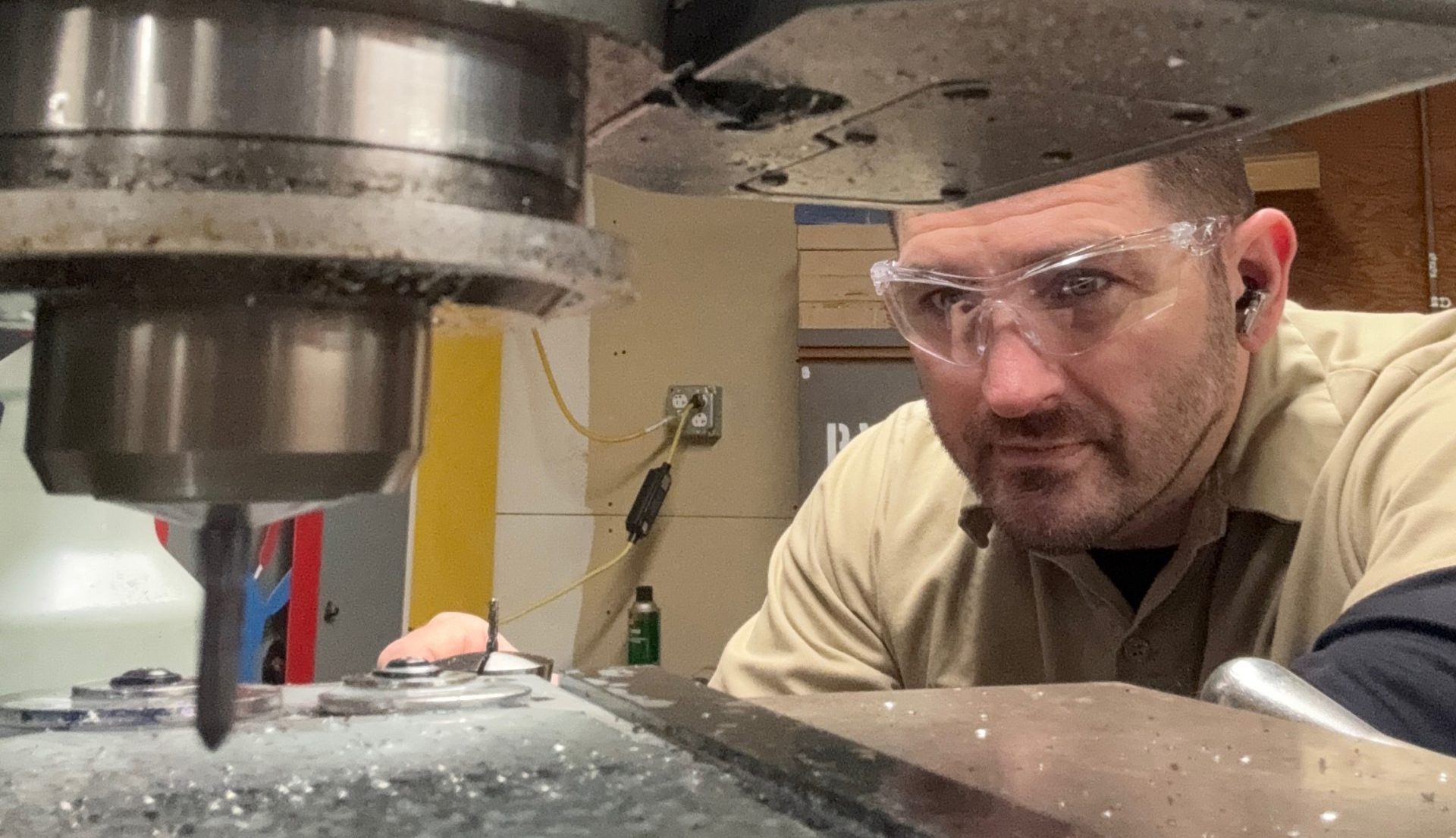 A man wearing safety glasses operates a large industrial milling machine, closely monitoring the drill bit cutting into metal.