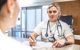 A Cleveland Clinic nurse wearing a stethoscope sits at a desk, writing on a form while speaking with another person in a hospital setting.