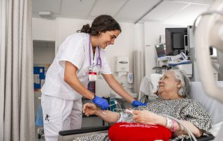 A nurse in white scrubs smiles while checking the IV of an older female patient lying in a hospital bed, as they share a warm interaction.
