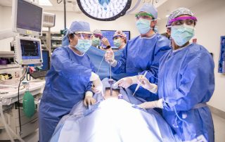 A team of surgeons in blue scrubs, masks, and protective eyewear perform a procedure under bright surgical lights in an operating room with monitoring equipment.