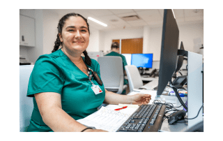 A woman healthcare worker in green scrubs smiles while sitting at a computer desk with paperwork and a keyboard in front of her.
