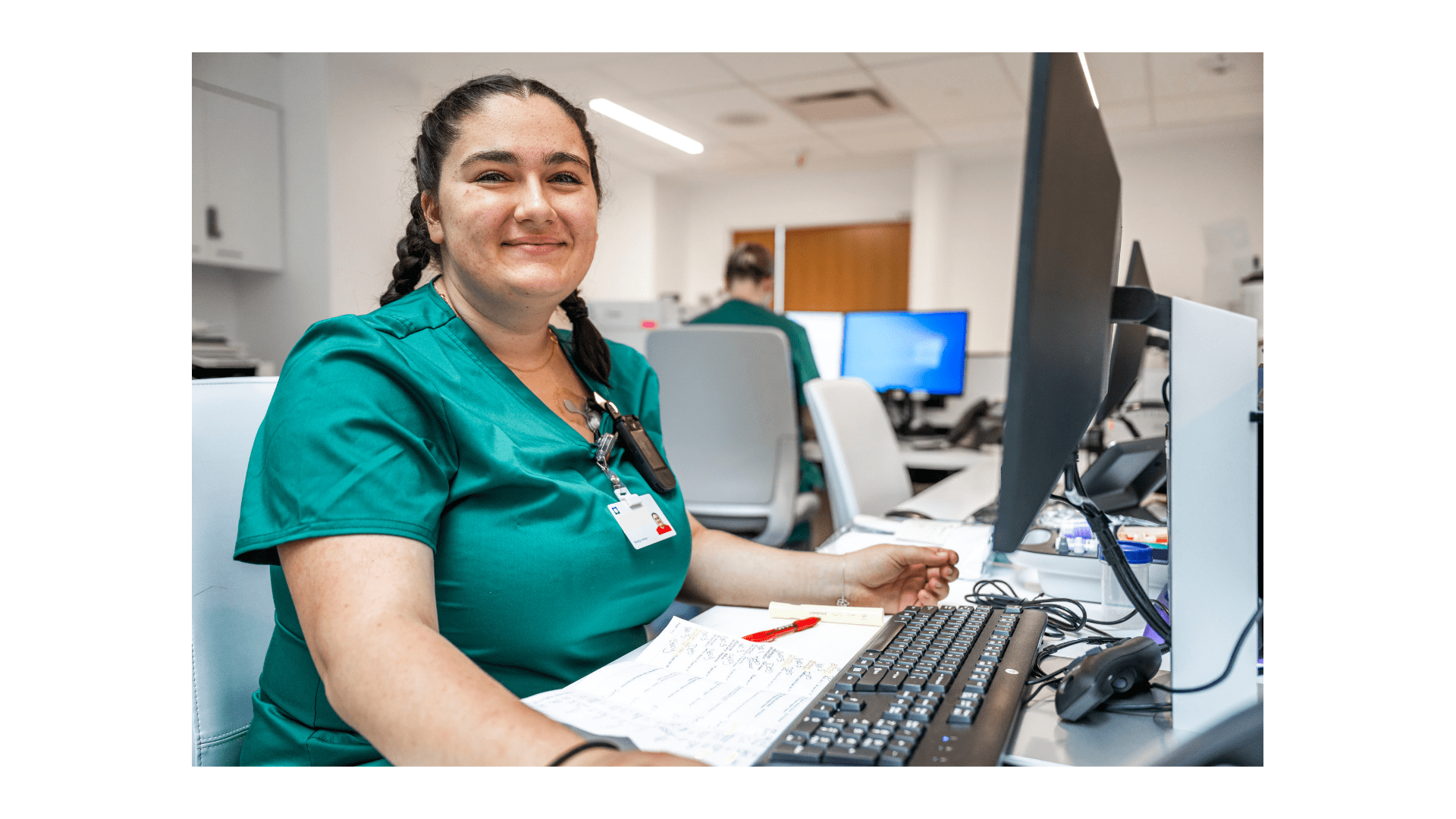 A woman healthcare worker in green scrubs smiles while sitting at a computer desk with paperwork and a keyboard in front of her.