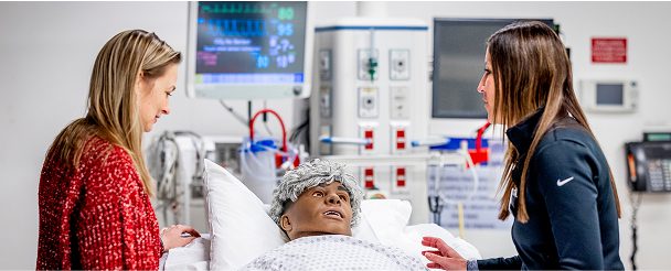Two women interact with a medical training mannequin lying in a hospital bed, with monitoring equipment visible in the background.