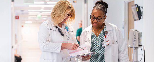 Two women healthcare professionals wearing white coats review documents together in a hospital hallway.