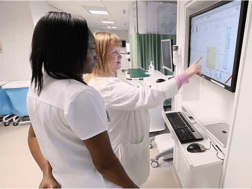 Two female nurses collaborating on a patient's case in front of a large wall-mounted computer monitor